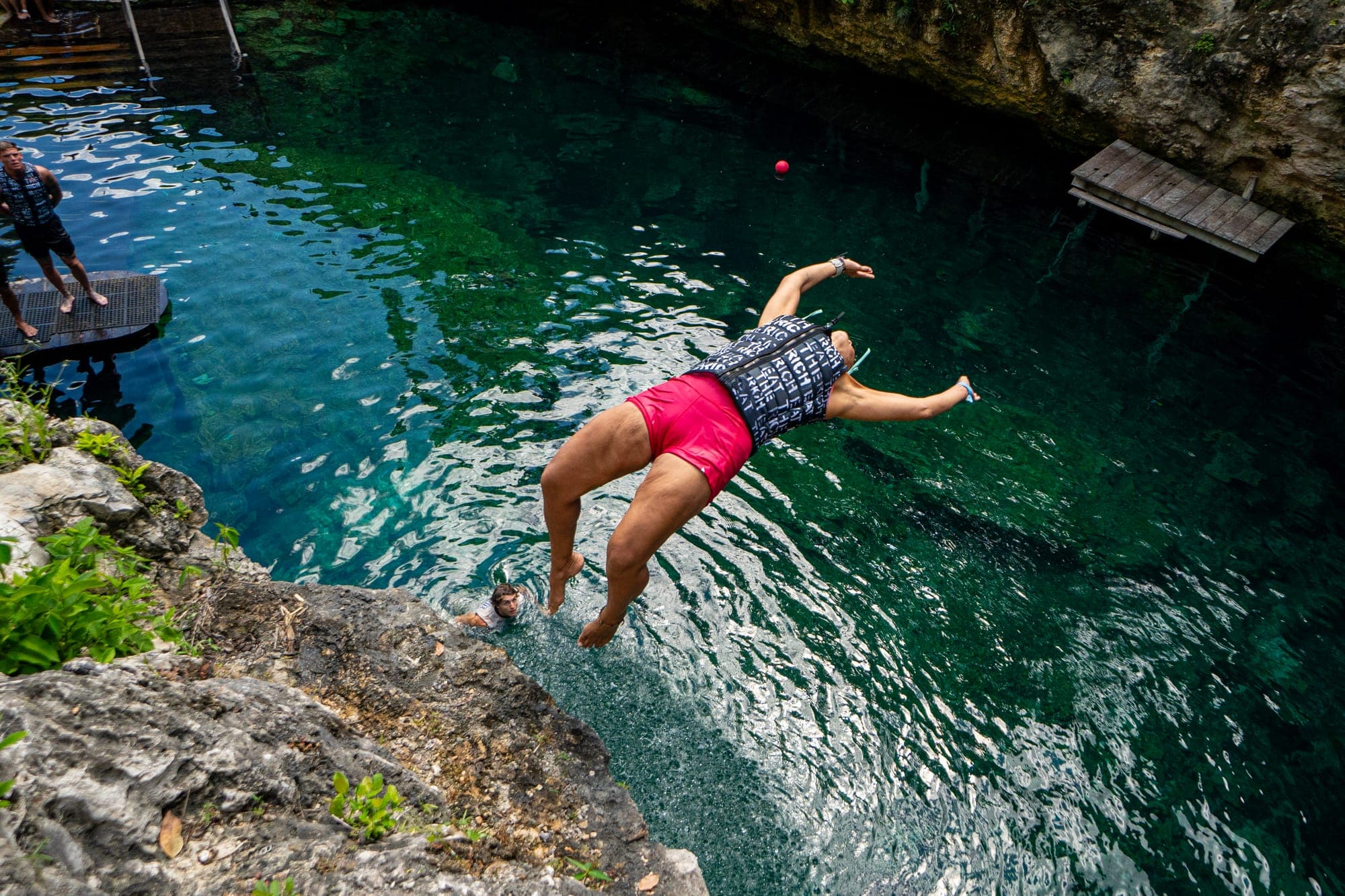 A jumper diving from a rocky ledge into clear turquoise cenote water